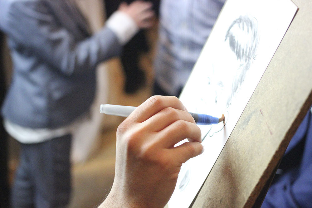 Un caricaturiste événementiel en train de dessiner sur le vif un invité lors d'un mariage. La caricature est réalisée avec du crayon, marqueur et de l'aquarelle.
